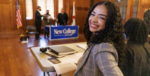 Smiling College age girl wearing a suit sits in beautiful wooden classroom where three teachers stand behind a table that has a large New College sign on it.